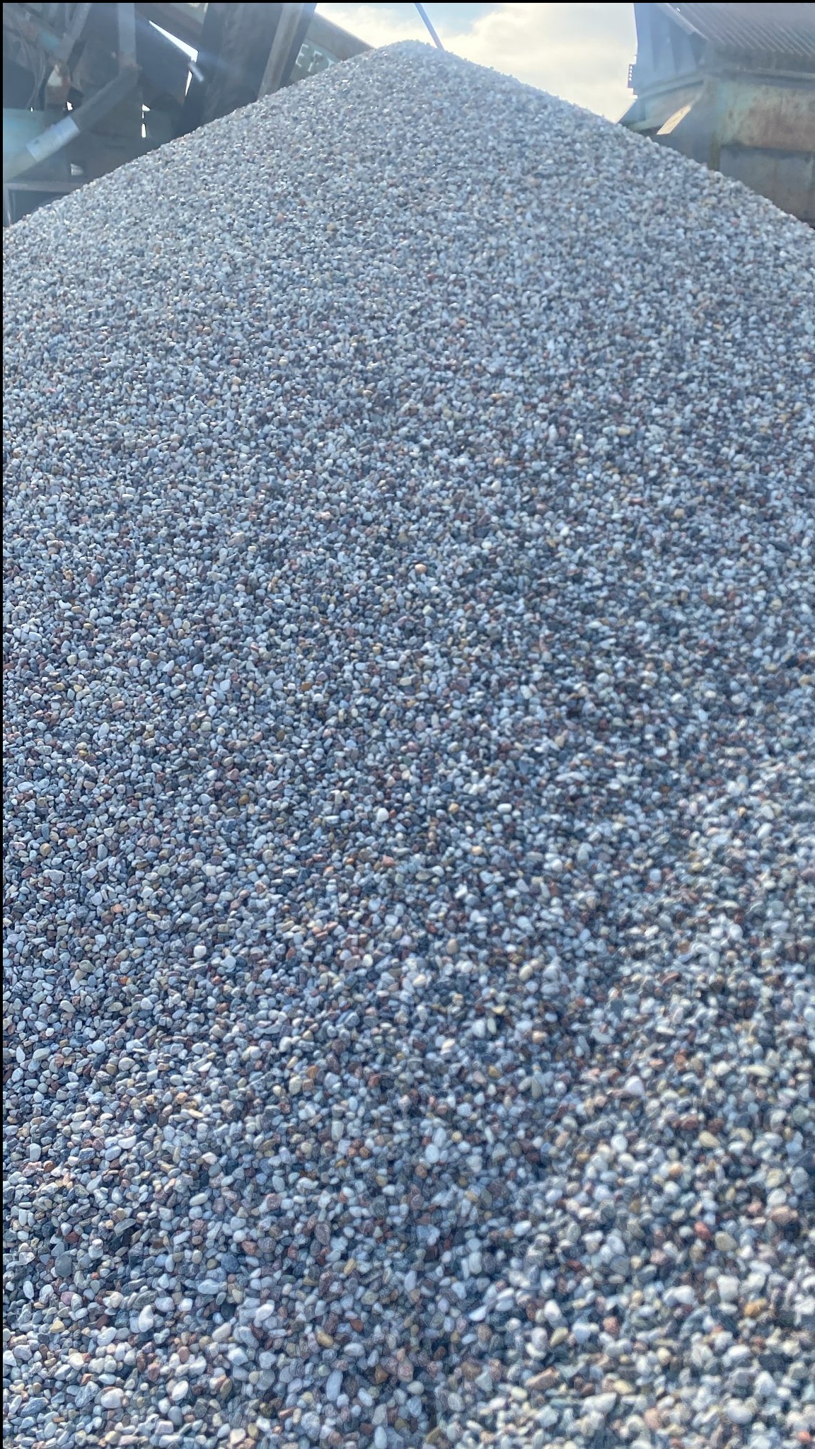 Góra drobnych kamieni o szaro-białej barwie, z widocznymi elementami maszyny do przesiewania w tle, oświetlona naturalnym światłem słonecznym.