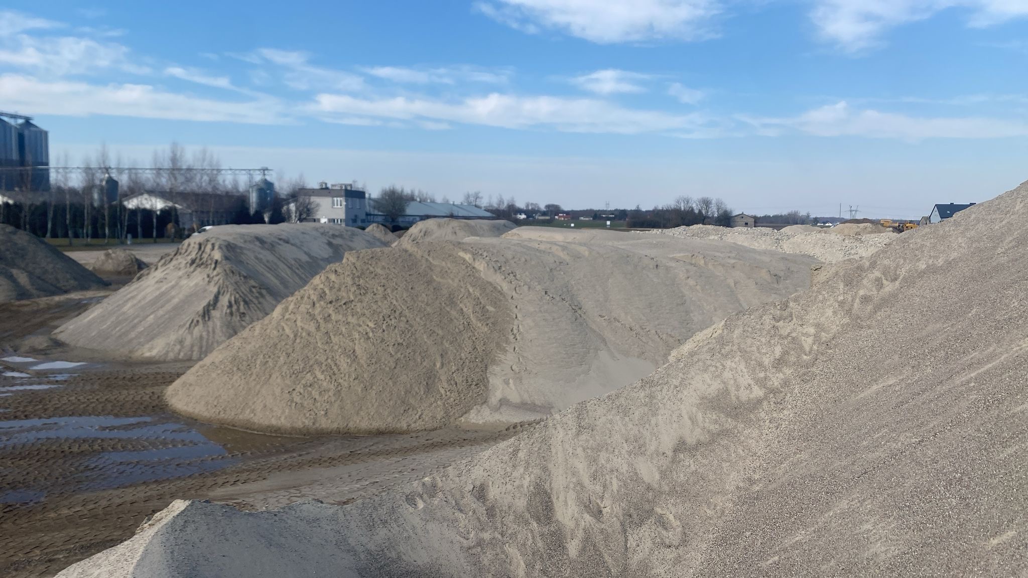 Spora pryzma jasnego kruszywa na placu budowy, widoczne ślady opon i kałuże, w tle budynki gospodarcze i błękitne niebo z chmurami.