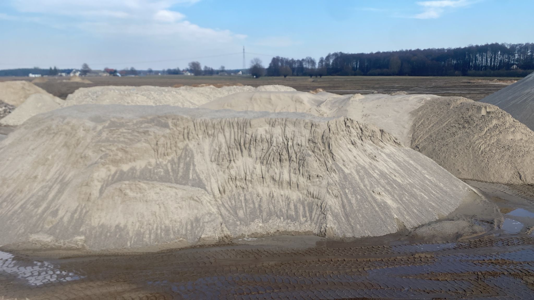 Kopalnia odkrywkowa z hałdami materiału sypkiego, widoczne ślady opon i kałuże, w tle pole i las.