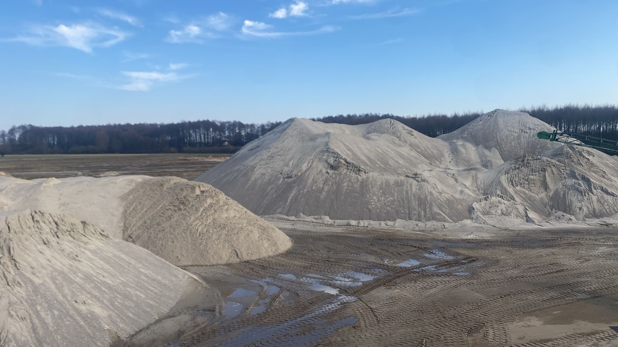 Kopalnia odkrywkowa z hałdami jasnego kruszywa, widoczne ślady opon i kałuże, w tle drzewa pod błękitnym niebem.
