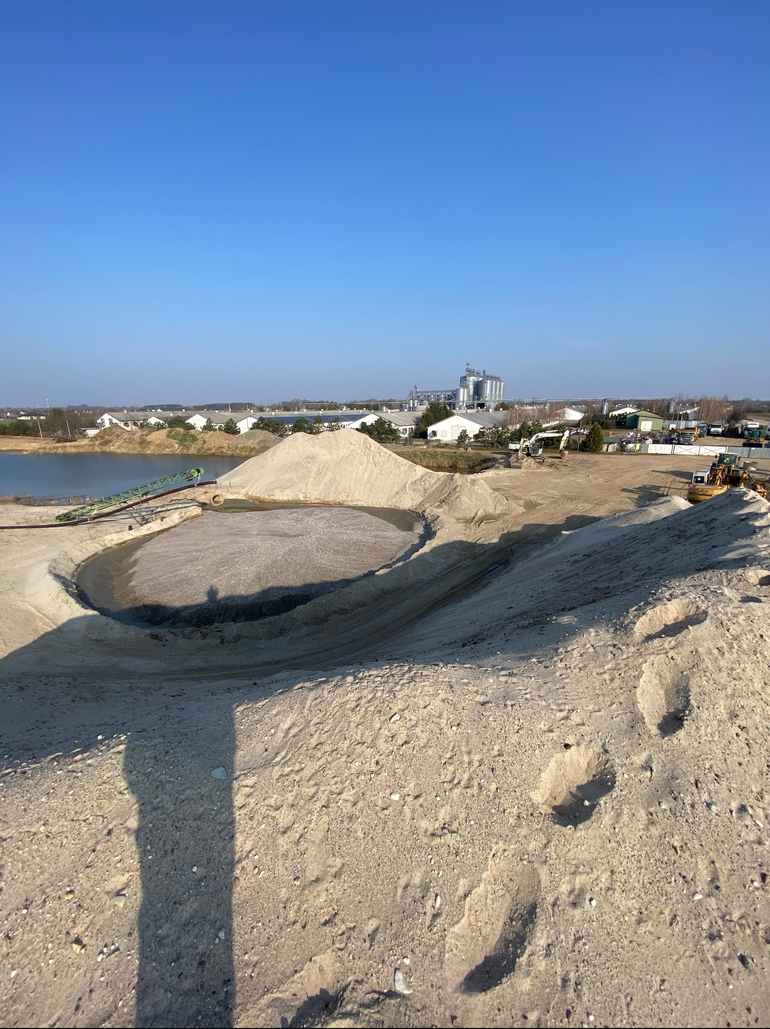 Kopalnia odkrywkowa z hałdami jasnego materiału, zbiornikiem wodnym i budynkami przemysłowymi w tle, widoczne ślady stóp na pierwszym planie.