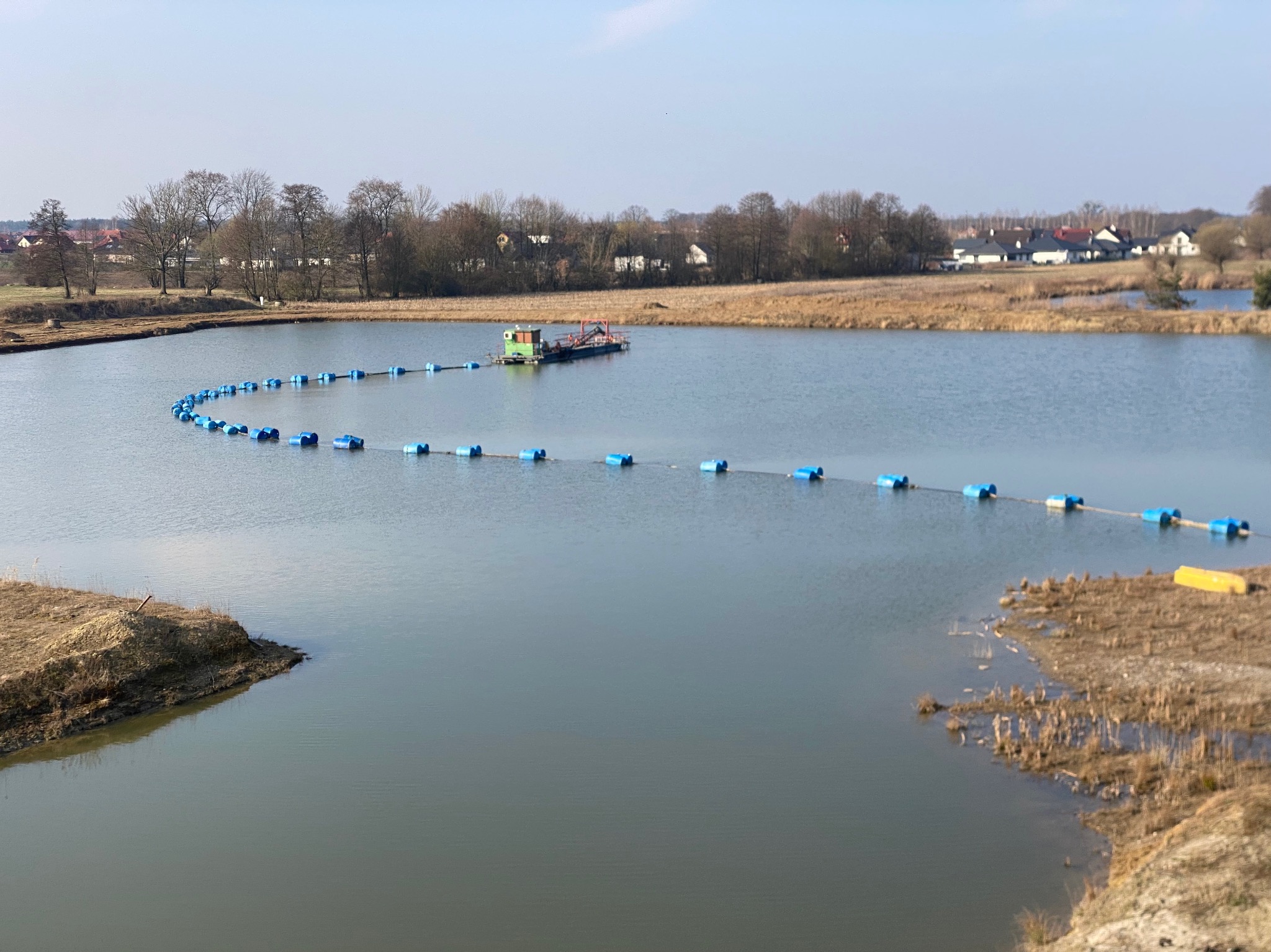 Pogłębianie dna zbiornika wodnego przy użyciu pływającej pogłębiarki z rurociągiem transportującym urobek, widoczne brzegi zbiornika z roślinnością i zabudowania w oddali.
