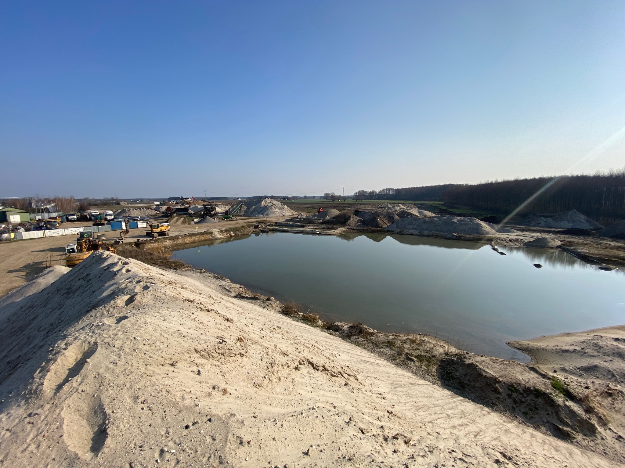 Kopalnia odkrywkowa z jeziorem w centrum, hałdy materiału, ciężki sprzęt budowlany w tle, błękitne niebo.