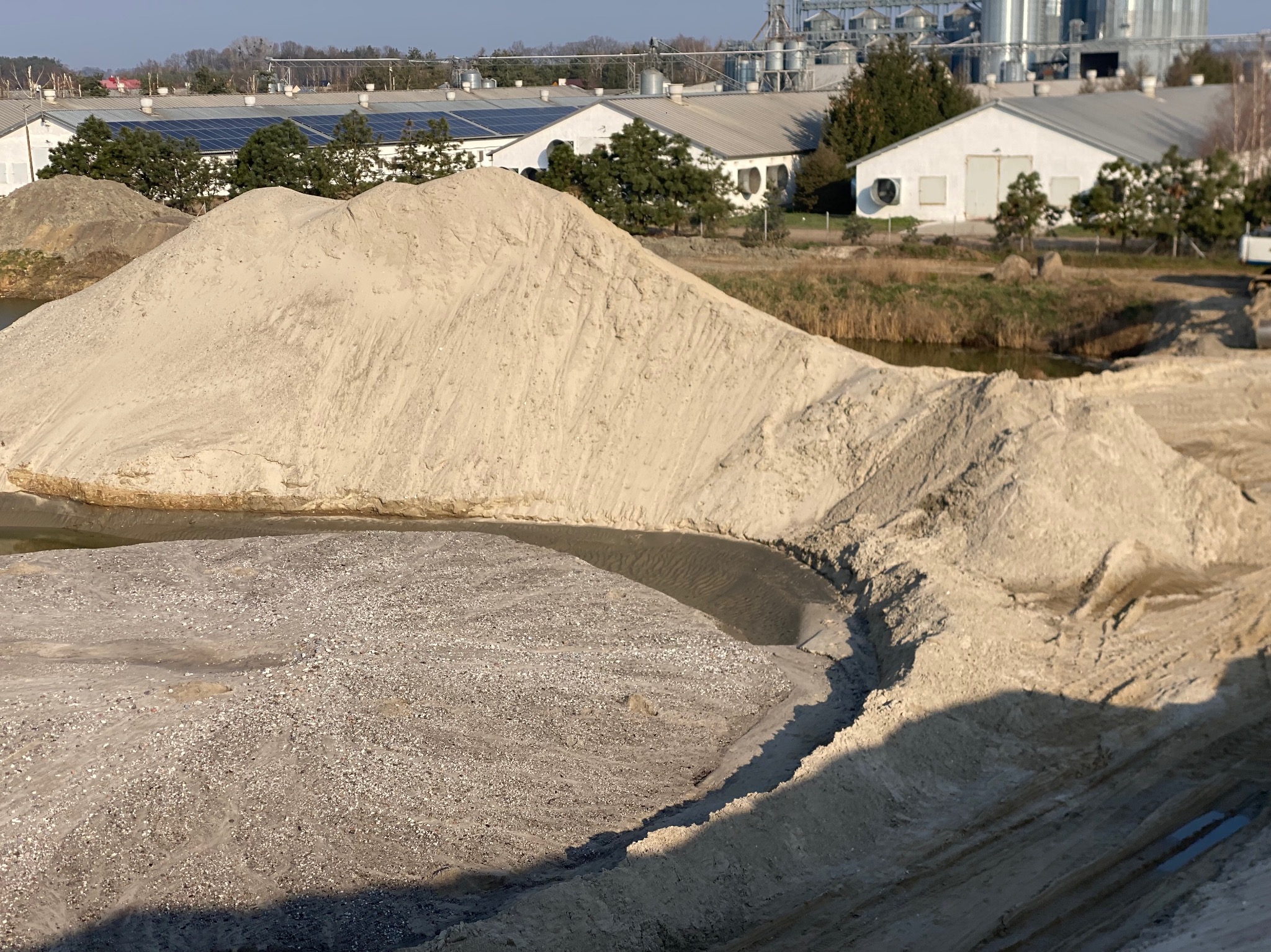 Duża pryzma jasnego materiału budowlanego na placu budowy, z budynkami gospodarczymi i silosami w tle.