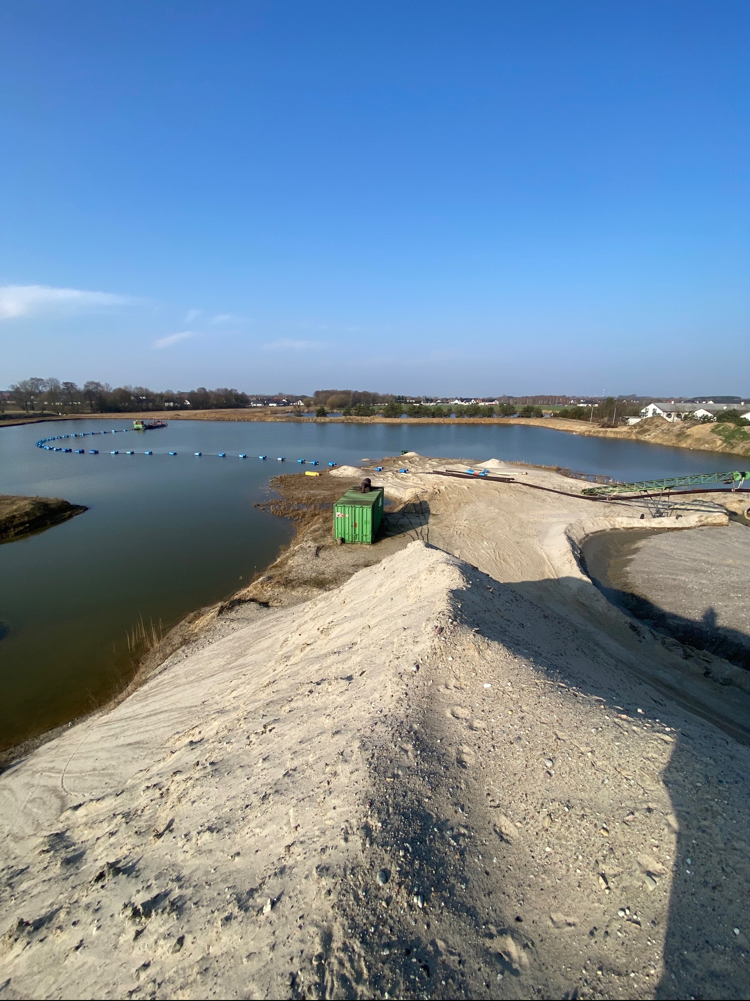 Kopalnia kruszywa z widokiem na zbiornik wodny, pływające rurociągi i zielony kontener w słoneczny dzień.
