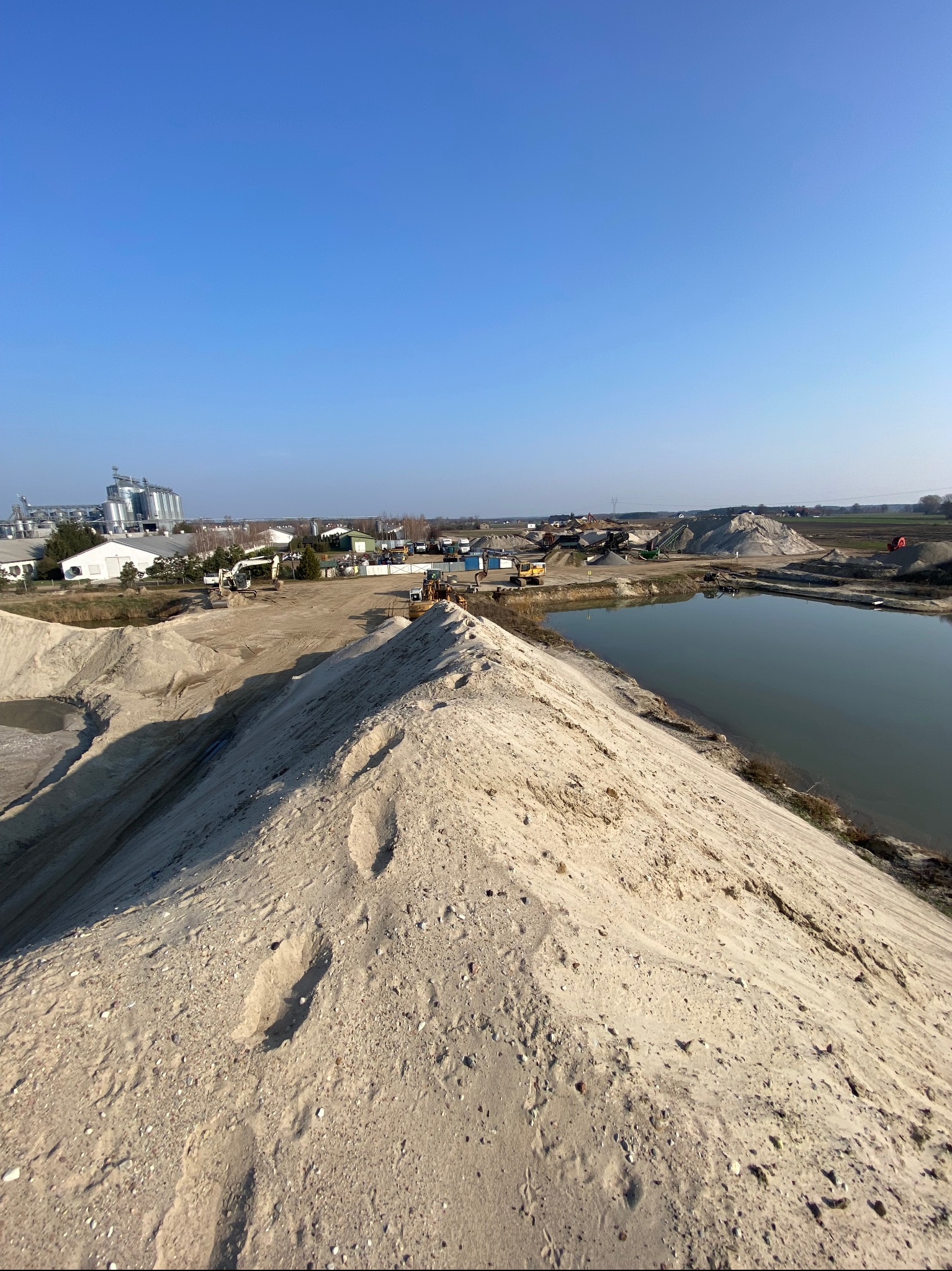 Widok z góry na hałdę materiału sypkiego z widocznymi śladami, obok zbiornik wodny, w tle zabudowania przemysłowe i sprzęt budowlany.