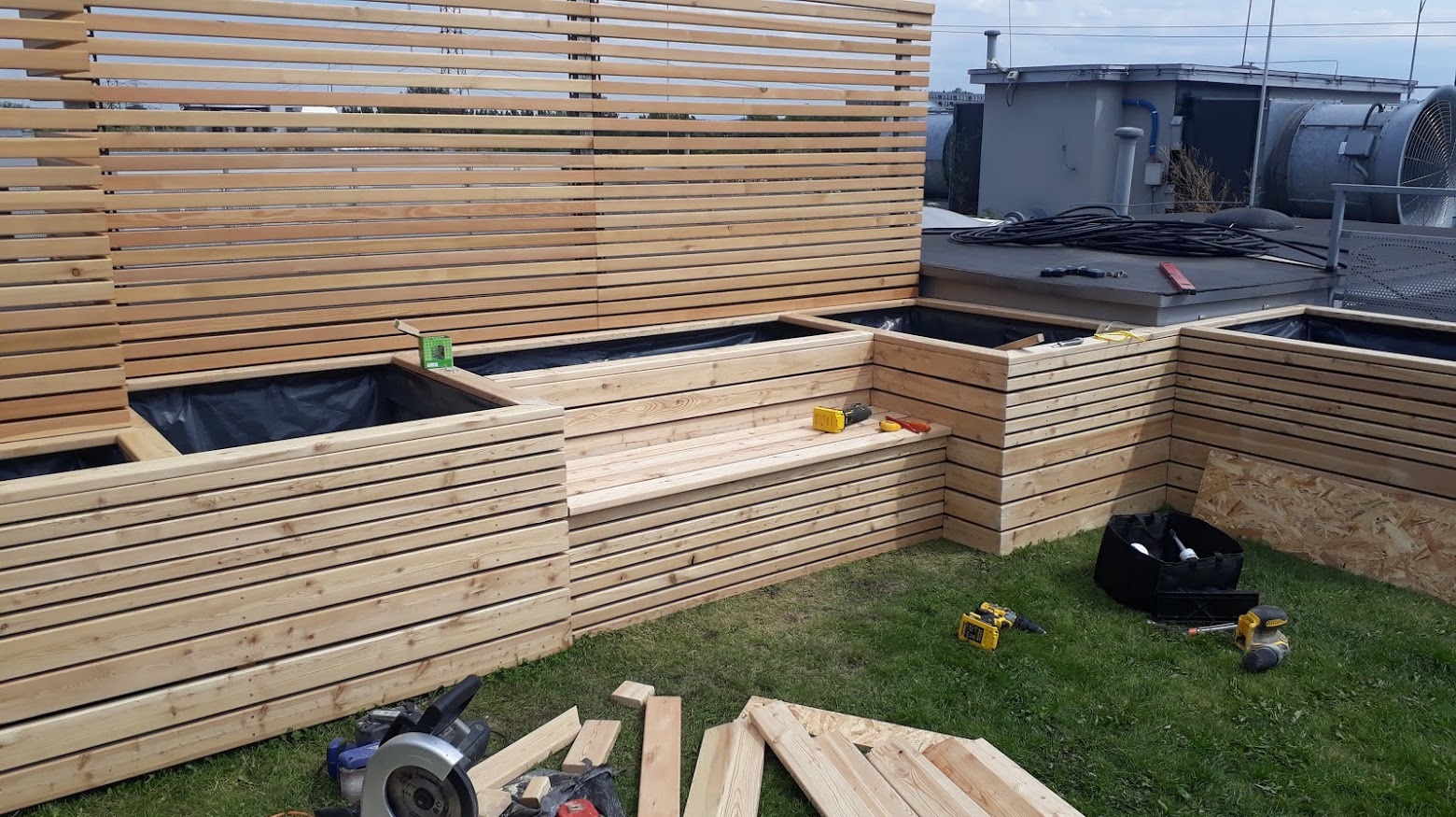 Drewniana konstrukcja z donicami i siedziskiem na dachu budynku, w trakcie montażu, z widocznymi narzędziami i elementami drewnianymi na trawie.