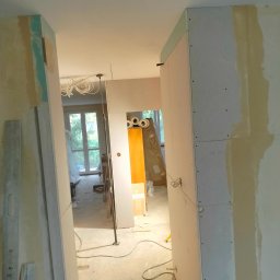 Remont marzeń - Wnętrze w trakcie remontu: widoczne płyty gipsowo-kartonowe na ścianach i sufitach, kable zwisające z sufitu, poziomica oparta o ścianę, okno z widokiem na zieleń w tle.