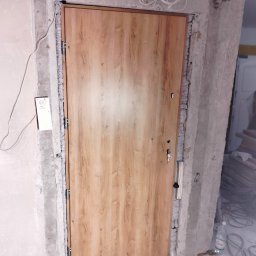 Remont marzeń - Montaż nowej, drewnopodobnej, brązowej drzwi wejściowych z widoczną pianką montażową w surowym otworze w ścianie, z wystającymi przewodami elektrycznymi u góry.