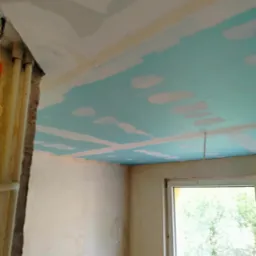 Niedokończone wnętrze: sufit z płyt gipsowo-kartonowych w trakcie szpachlowania, ściany w stanie surowym, widoczne instalacje przy ścianie, okno z widokiem na zieleń.