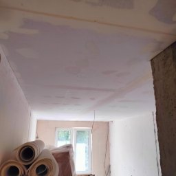 Remont marzeń - Wnętrze w trakcie remontu: sufit i ściany po nałożeniu gładzi, widoczne rolki papieru malarskiego, okno w tle.