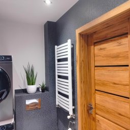 Remont marzeń - Nowoczesna łazienka z szarymi mozaikowymi płytkami, drewnianymi drzwiami, białym grzejnikiem drabinkowym i pralko-suszarką.