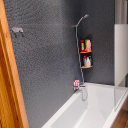 Remont marzeń - Nowoczesna łazienka z ciemnoszarą mozaiką na ścianie, białą wanną, szklanym parawanem nawannowym i drewnianymi elementami wykończeniowymi.