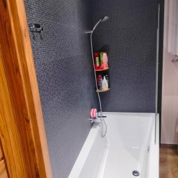 Remont marzeń - Nowoczesna łazienka z wanną, ściany wyłożone drobną szarą mozaiką, drewniane elementy wykończeniowe i półki z kosmetykami.