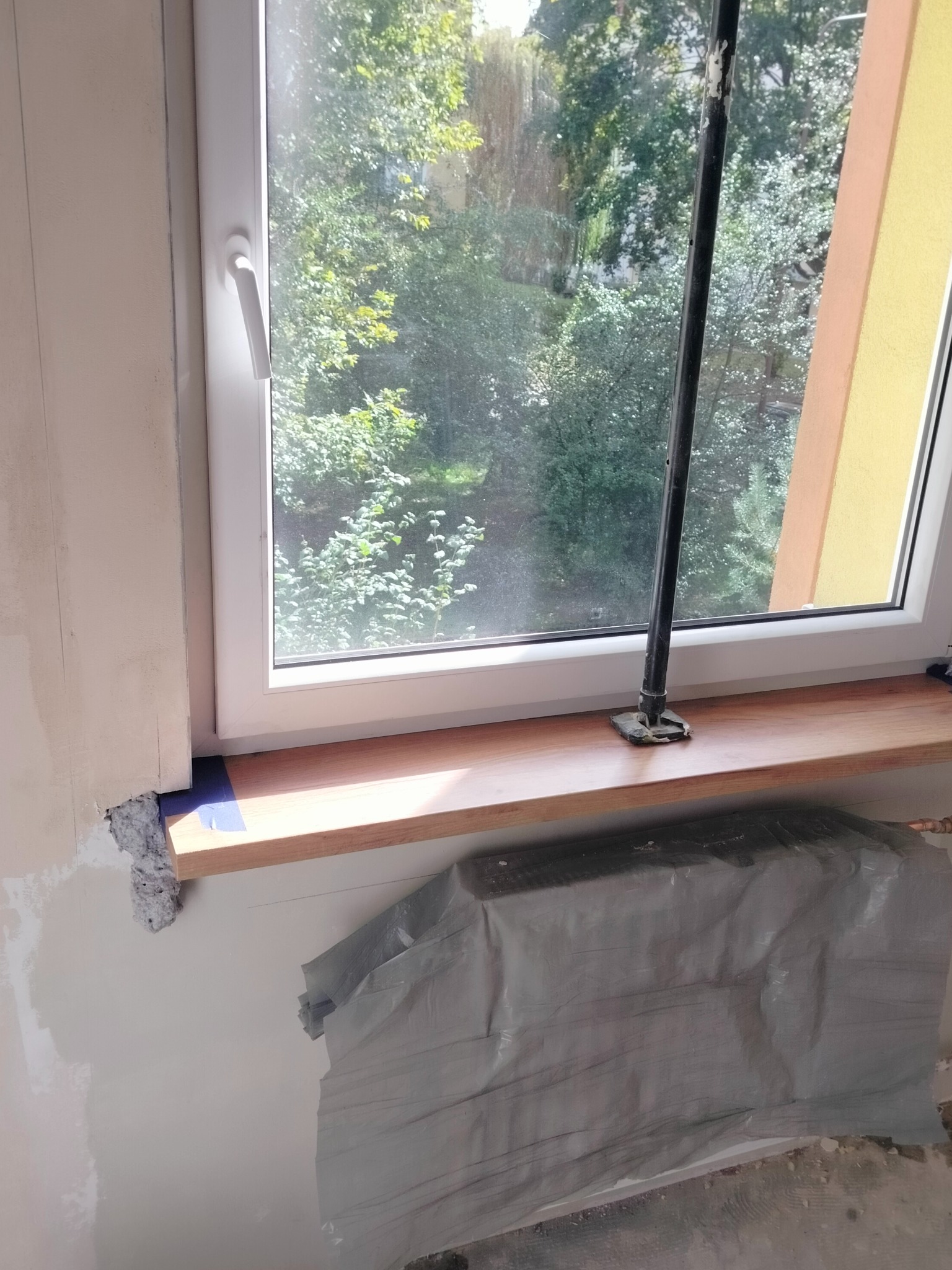 Przygotowanie do prac wykończeniowych przy oknie: zabezpieczony grzejnik folią, świeżo położona gładź na ścianie, drewniany parapet z niebieską taśmą malarską, podpora teleskopowa oparta o parapet.