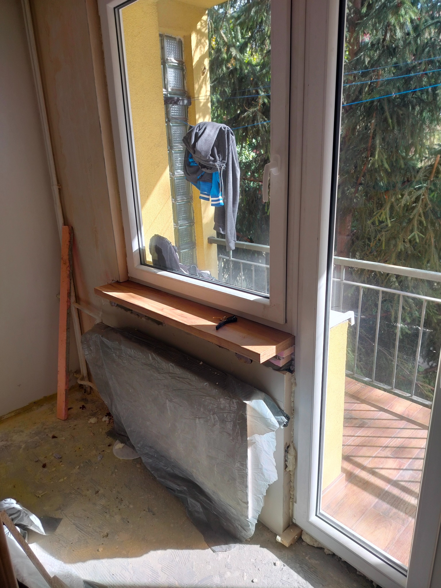 Montaż parapetu wewnętrznego z drewna, okno balkonowe z widokiem na balkon i zieleń, zabezpieczony folią grzejnik centralnego ogrzewania, widoczne ślady prac remontowych.