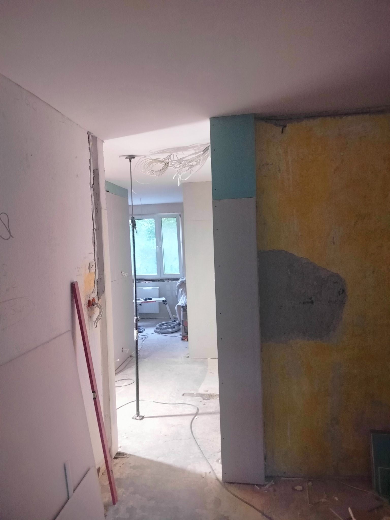 Wnętrze w trakcie remontu: widoczne płyty gipsowo-kartonowe, fragment starej ściany z resztkami farby, wystające przewody elektryczne i poziomica oparta o ścianę.