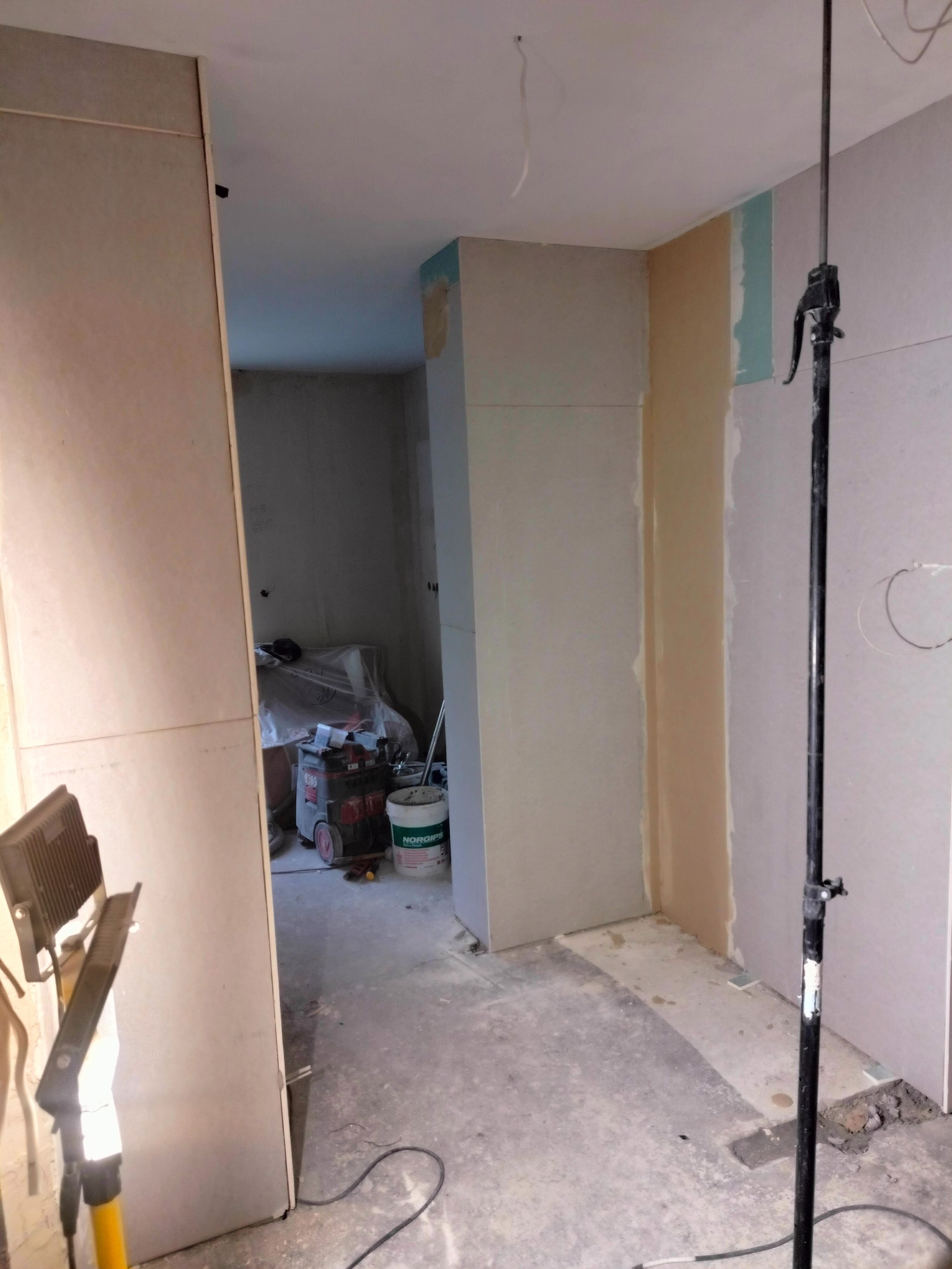 Wnętrze w trakcie prac wykończeniowych: widoczne płyty gipsowo-kartonowe na ścianach, szpachlowane łączenia, lampa budowlana na statywie oraz wiadro z masą szpachlową w tle.