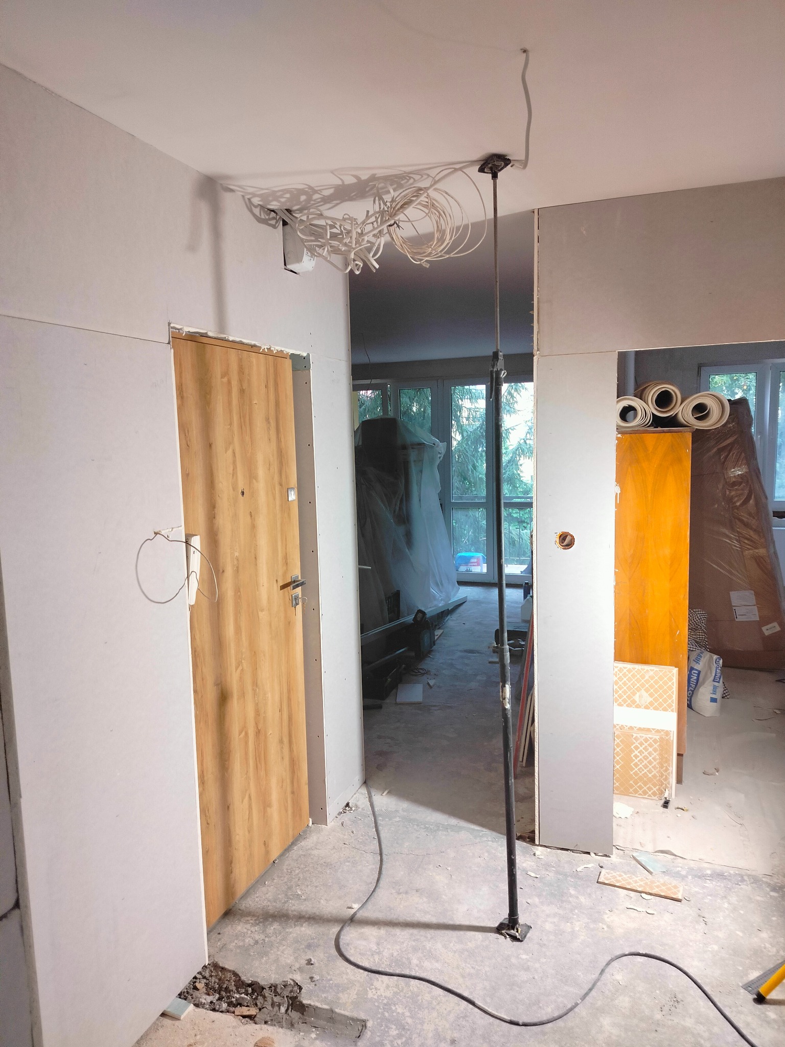 Wnętrze w trakcie remontu: zamontowane płyty gipsowo-kartonowe, nowa drewnopodobna ościeżnica z drzwiami, wystające przewody elektryczne na suficie, podpora pionowa, podłoga z fragmentami gruzu.