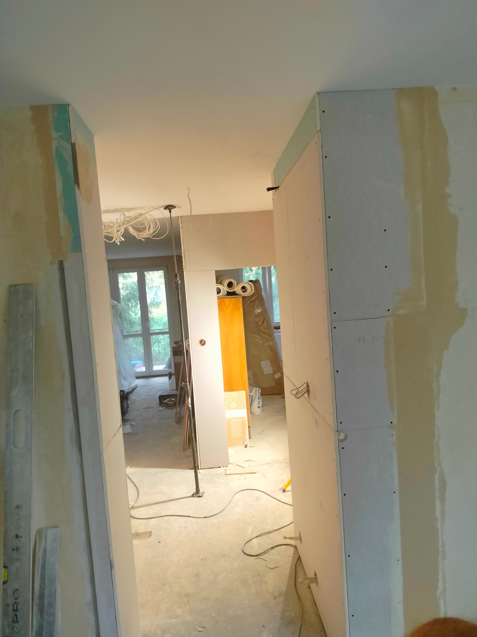Wnętrze w trakcie remontu: widoczne płyty gipsowo-kartonowe na ścianach i sufitach, kable zwisające z sufitu, poziomica oparta o ścianę, okno z widokiem na zieleń w tle.