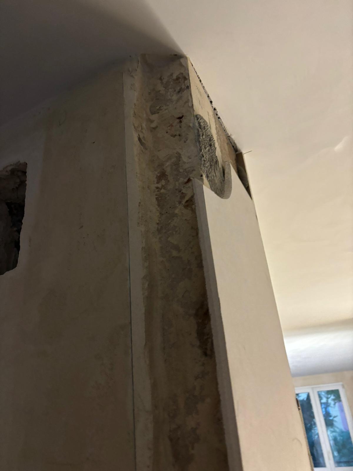 Narożnik ściany w trakcie przygotowania do wykończenia, widoczne surowe tynki i fragment płyty gipsowej, okno w tle.