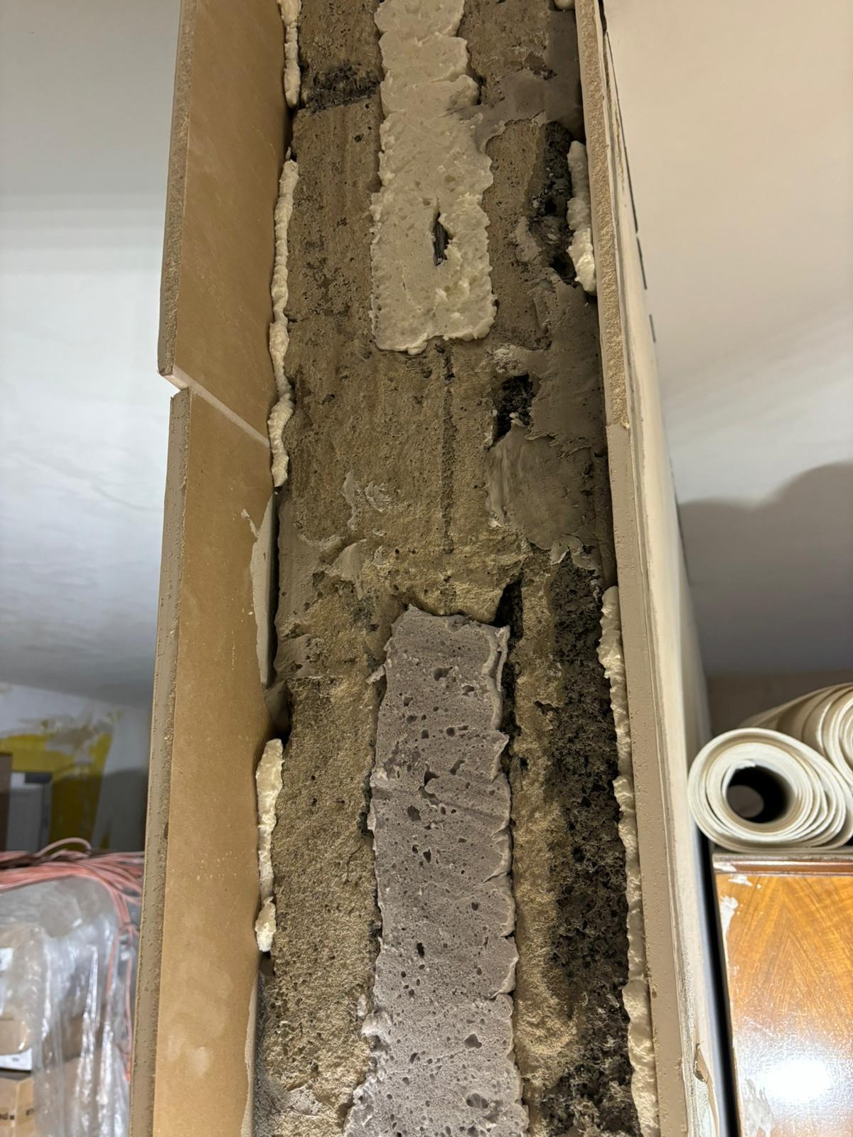 Fragment ściany z odsłoniętą strukturą, widoczne warstwy: stara zaprawa, pianka montażowa, płyta karton-gips z widocznym pęknięciem na łączeniu, w tle rolki tapet.
