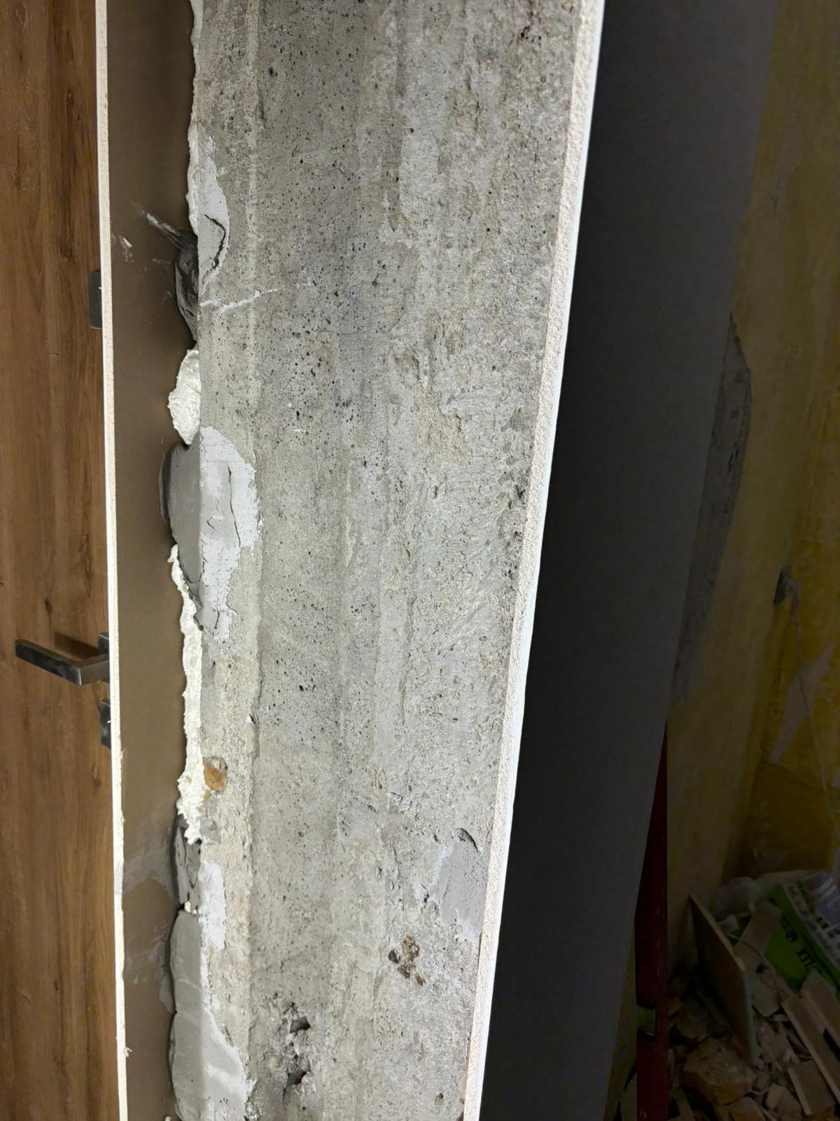 Niedokończone wykończenie ściany z widocznym betonem, częściowo pokryte gładzią, z fragmentem ościeżnicy drzwi po lewej stronie i resztkami tapety w tle po prawej.