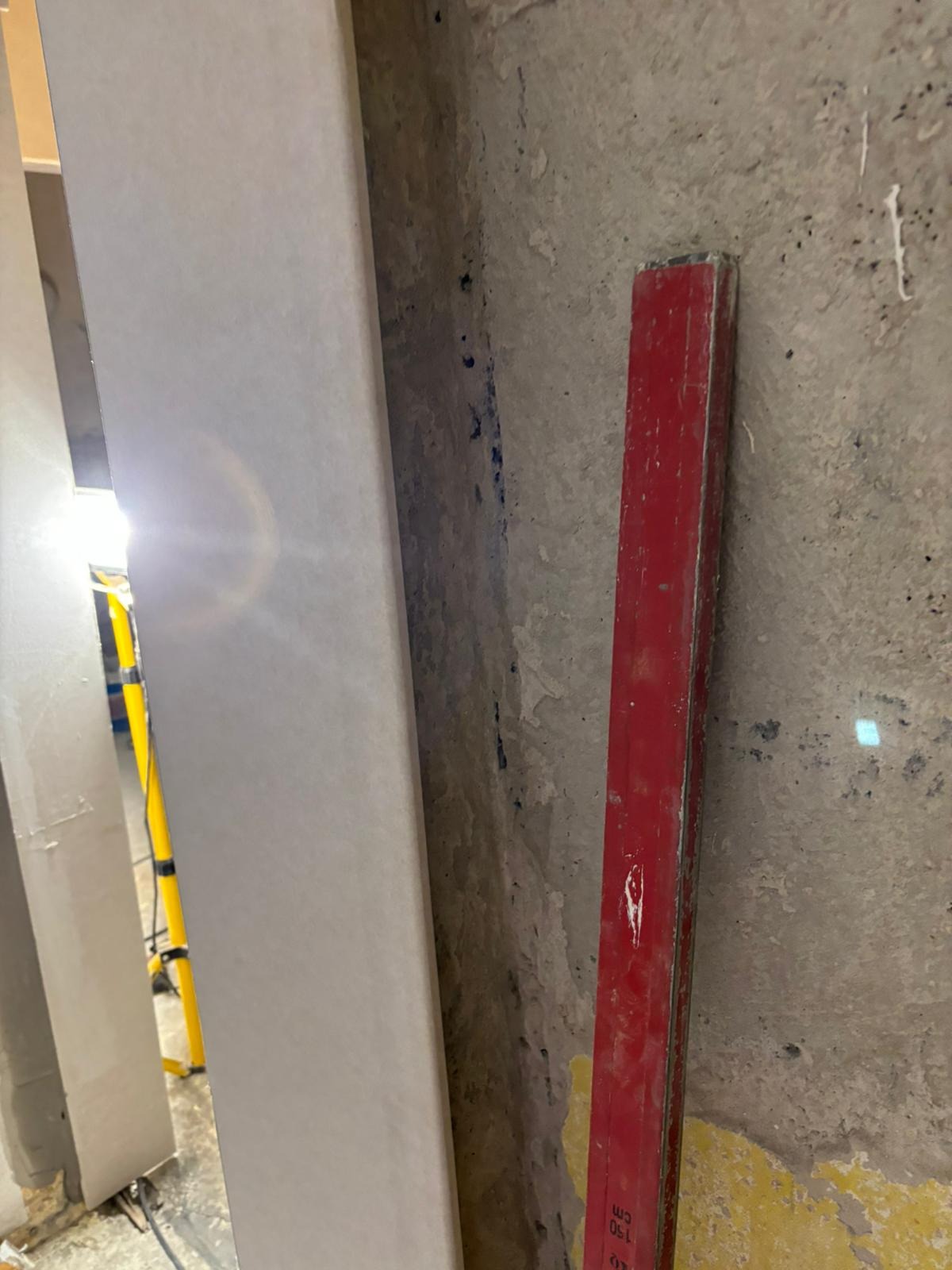 Ściana w trakcie remontu z widoczną łatą gipsową, starą czerwoną poziomicą i lampą budowlaną w tle.