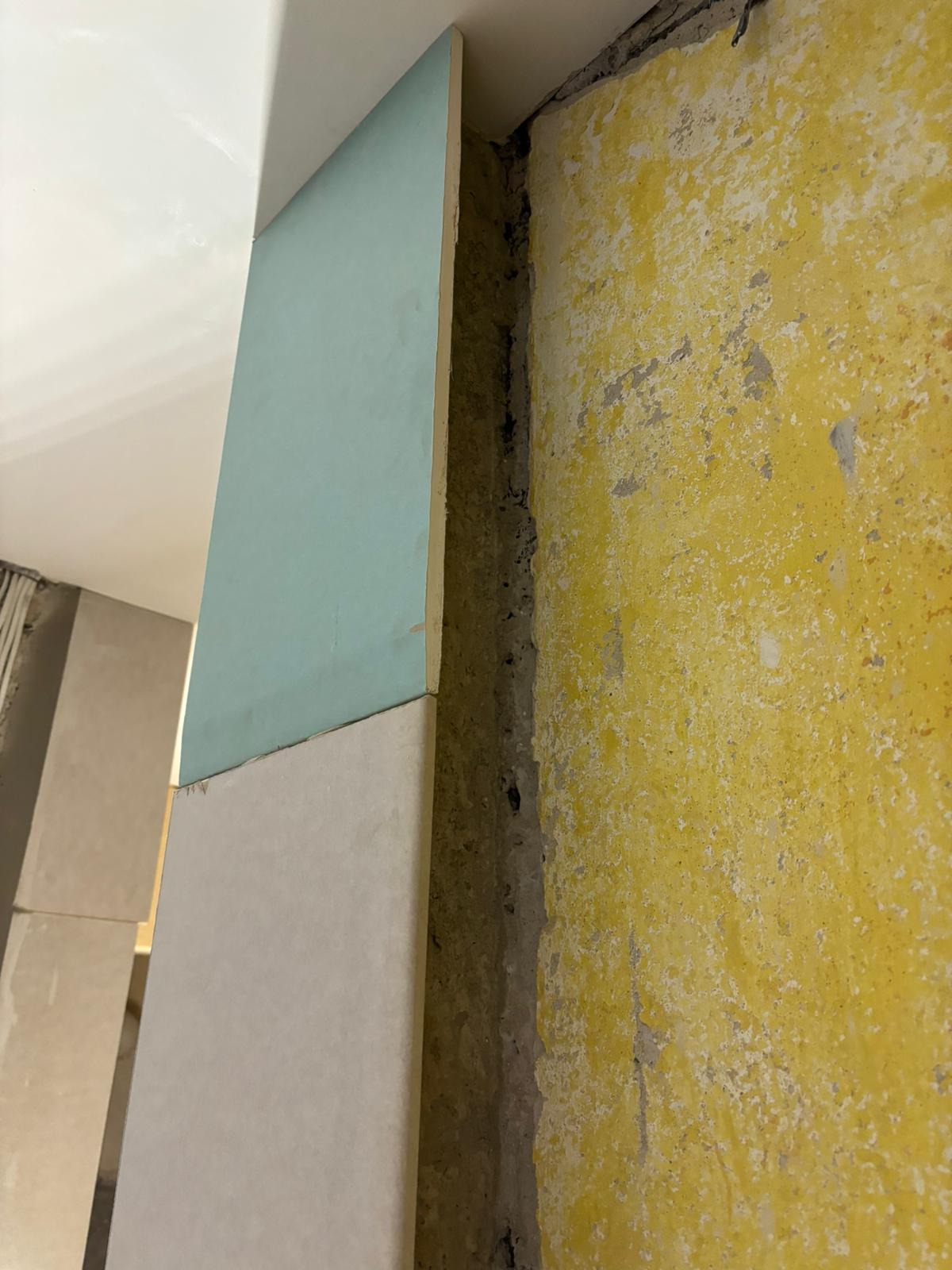 Fragment ściany z żółtą, łuszczącą się farbą, obok której zamontowano płytę gipsowo-kartonową, częściowo pomalowaną na jasny, pastelowy odcień niebieskiego, z widoczną szczeliną pomiędzy płytą...