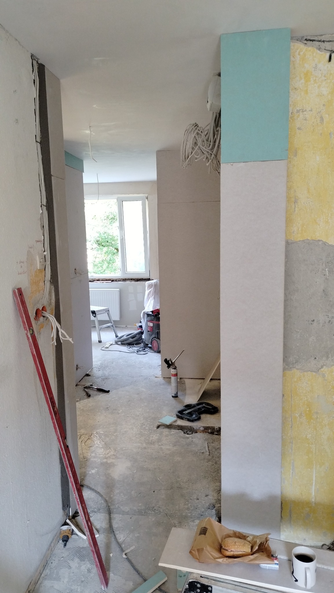 Wnętrze w trakcie prac, widoczne płyty kartonowo-gipsowe, przewody elektryczne wystające ze ściany, poziomica oparta o ścianę, narzędzia i posiłek na stole, okno w tle.