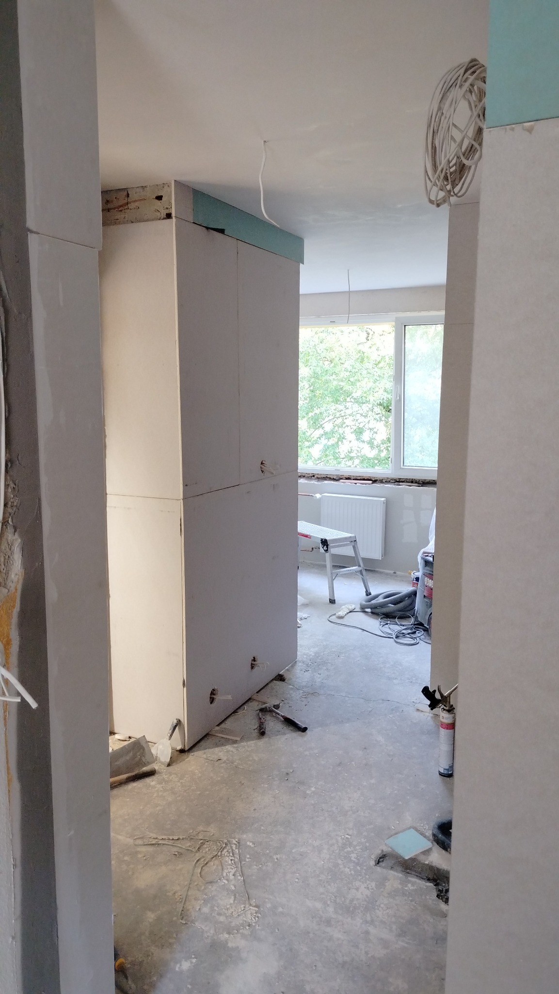 Wnętrze w trakcie prac wykończeniowych: widok na pokój z surową podłogą, częściowo zabudowaną ścianą z płyt gipsowo-kartonowych, narzędzia budowlane na podłodze i okno w tle.