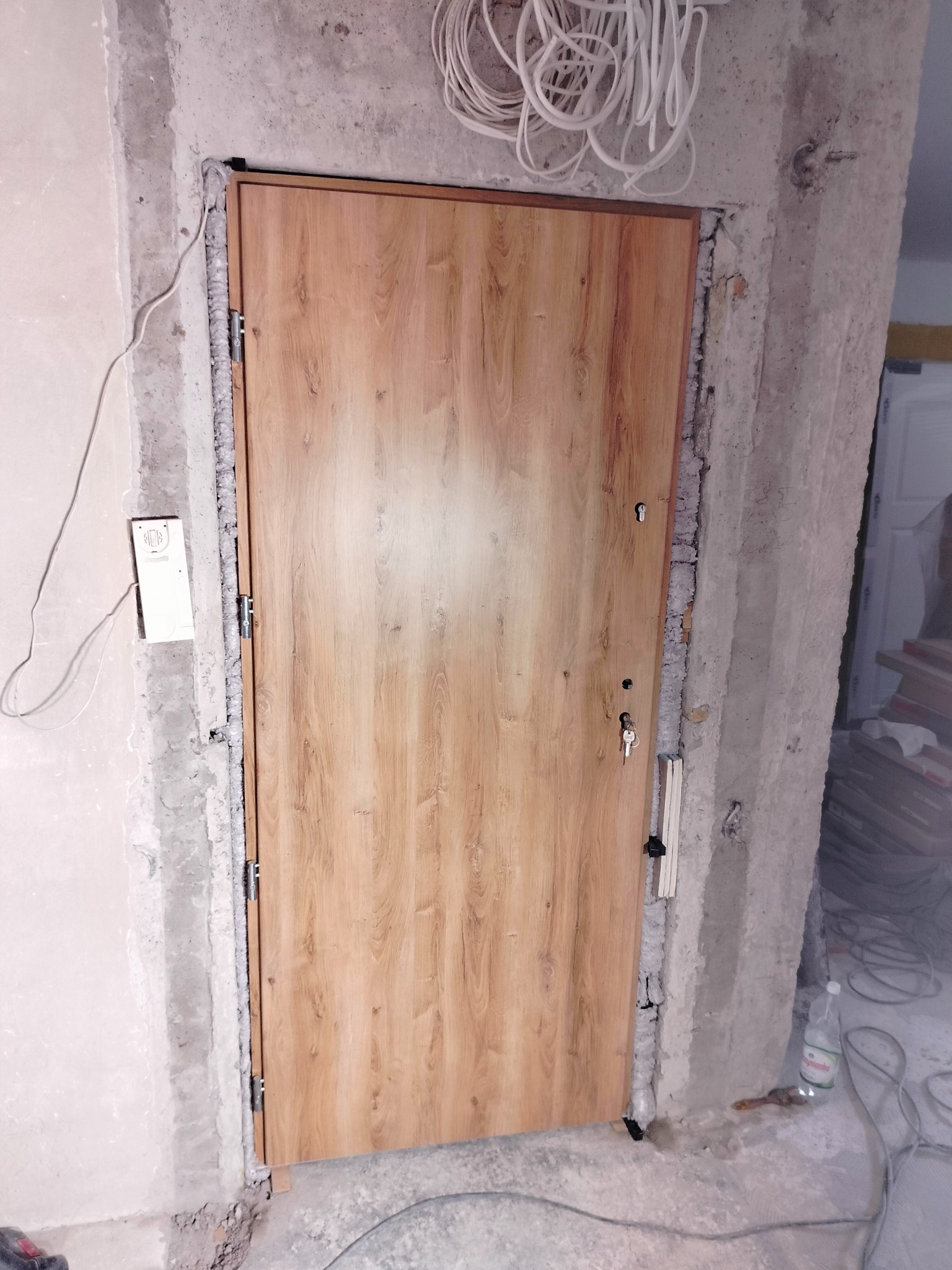 Montaż nowej, drewnopodobnej, brązowej drzwi wejściowych z widoczną pianką montażową w surowym otworze w ścianie, z wystającymi przewodami elektrycznymi u góry.