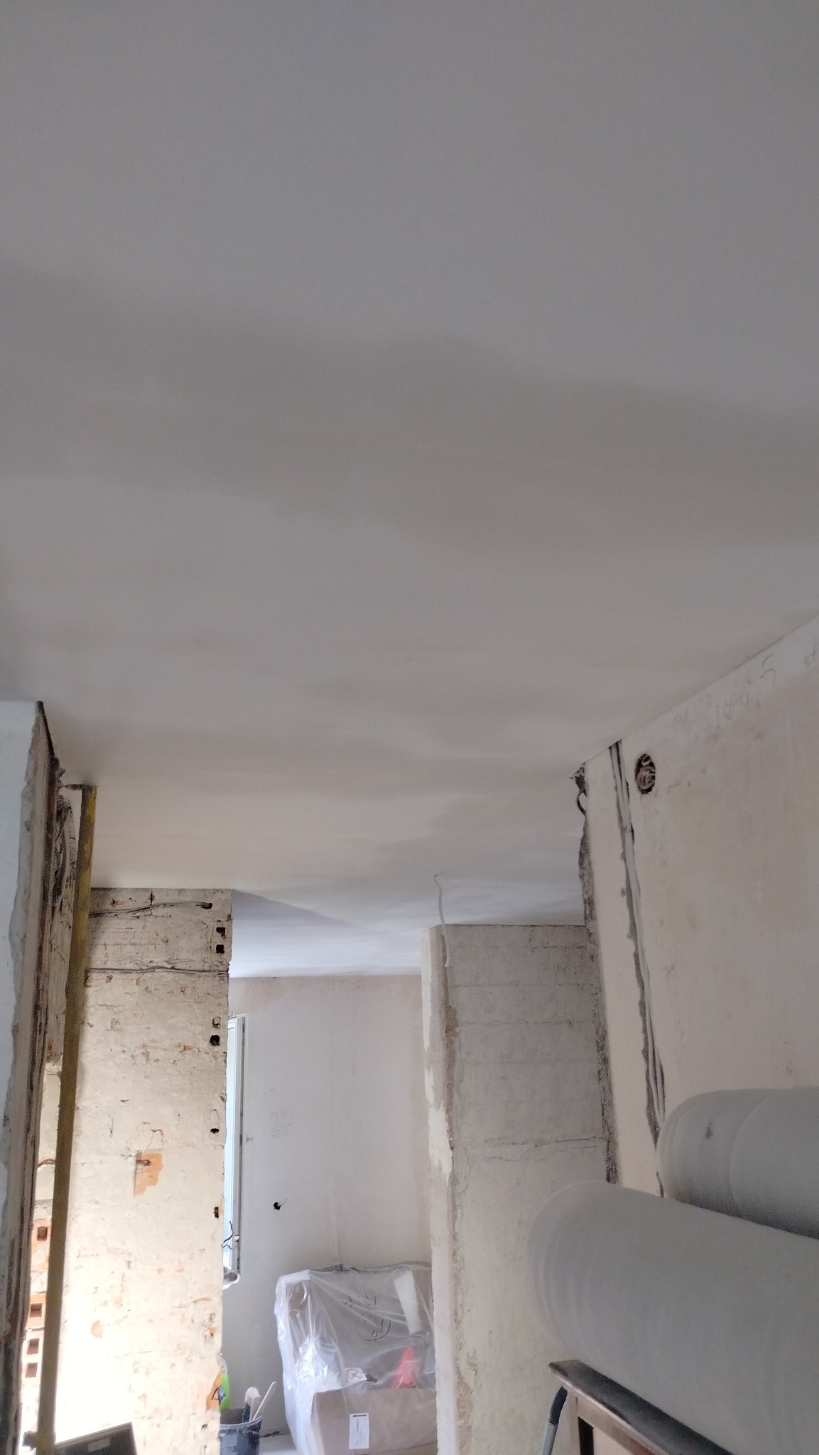 Wnętrze w trakcie remontu, widoczne ściany w stanie surowym, częściowo pokryte gładzią, odsłonięta cegła, materiały budowlane zabezpieczone folią w tle.