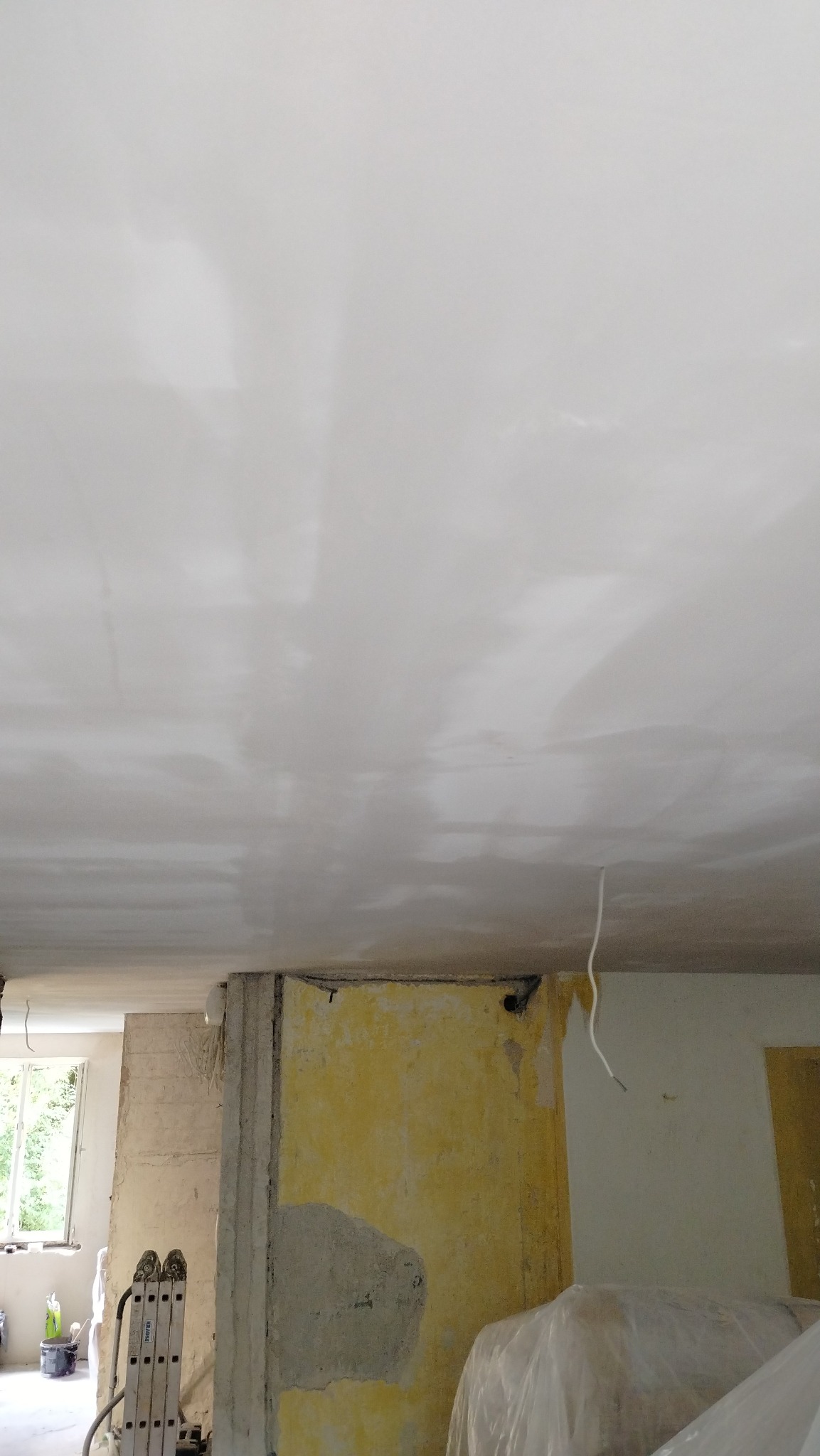 Niedokończone wnętrze w trakcie remontu: sufit po nałożeniu gładzi, widoczne fragmenty ścian w różnych kolorach i fakturach, drabina oparta o ścianę, okno w tle.