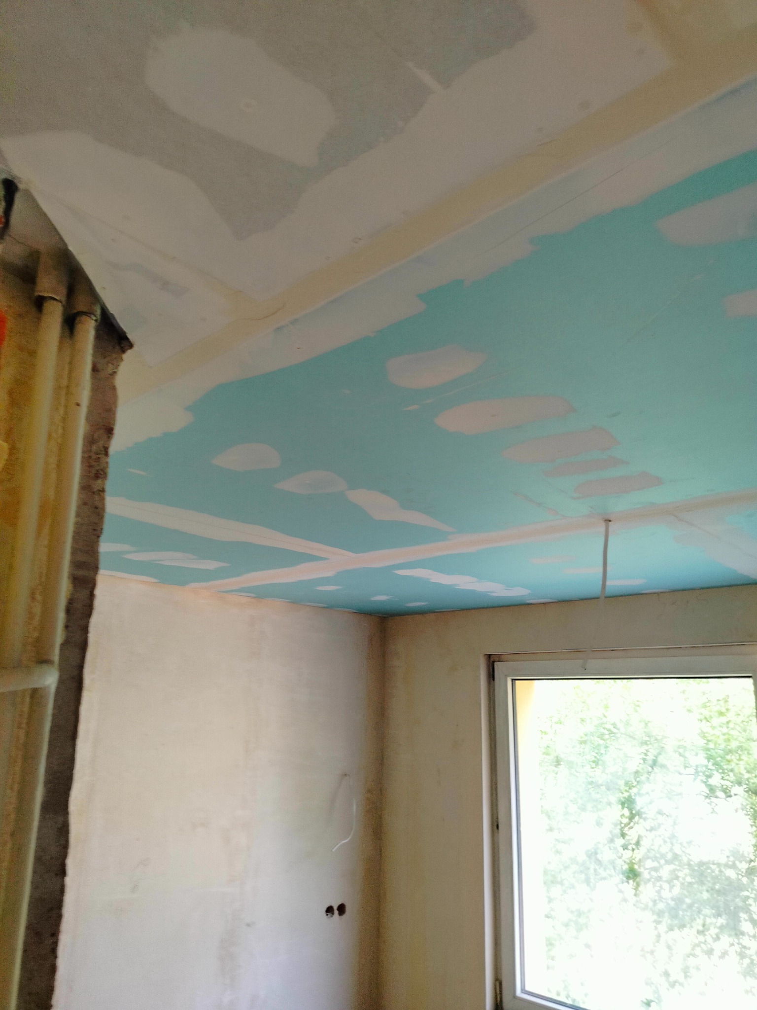 Niedokończone wnętrze: sufit z płyt gipsowo-kartonowych w trakcie szpachlowania, ściany w stanie surowym, widoczne instalacje przy ścianie, okno z widokiem na zieleń.