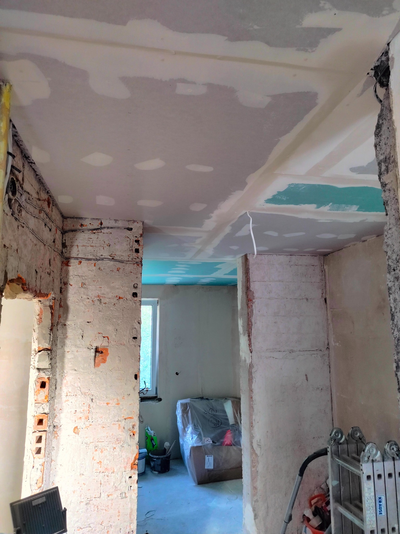 Wnętrze w trakcie prac przygotowawczych do wykończenia, widoczne surowe ściany z cegły częściowo pokryte gładzią, fragment sufitu z nałożoną warstwą gładzi, przewody elektryczne i drabina aluminiowa.