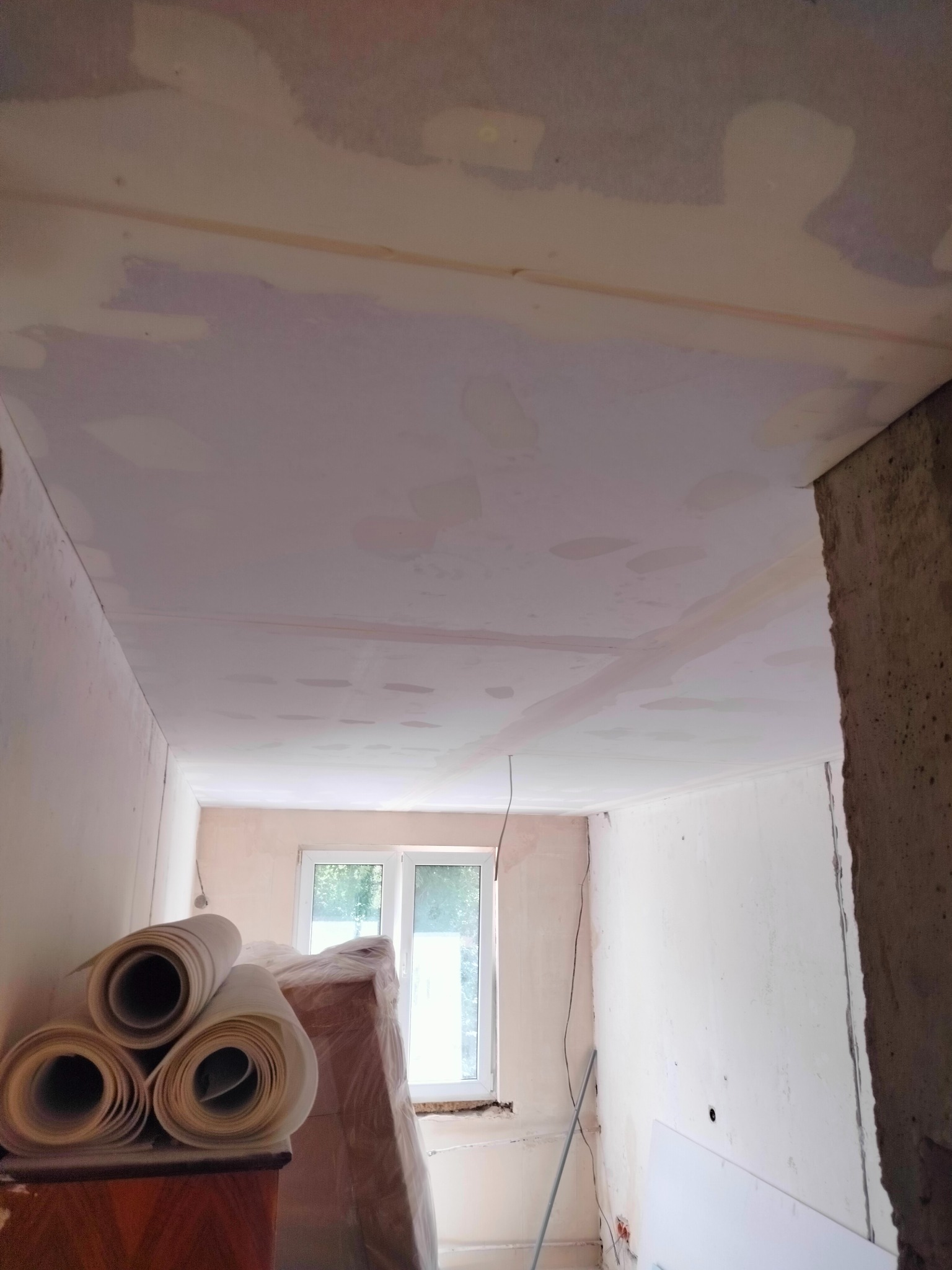 Wnętrze w trakcie remontu: sufit i ściany po nałożeniu gładzi, widoczne rolki papieru malarskiego, okno w tle.
