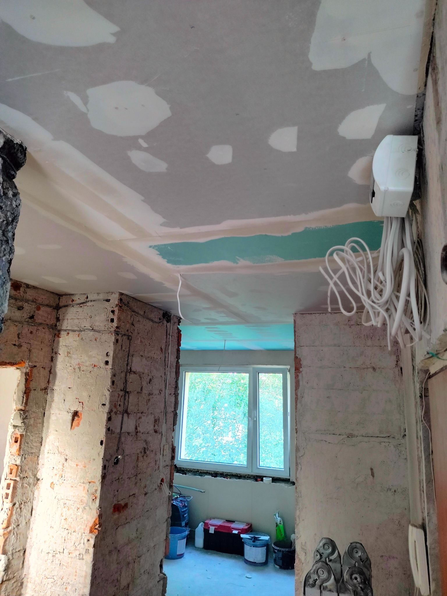 Wnętrze w trakcie prac przygotowawczych do wykończenia: surowa cegła, częściowo położona gładź na suficie, wystające przewody elektryczne z puszki, widok na okno z zielenią na zewnątrz.