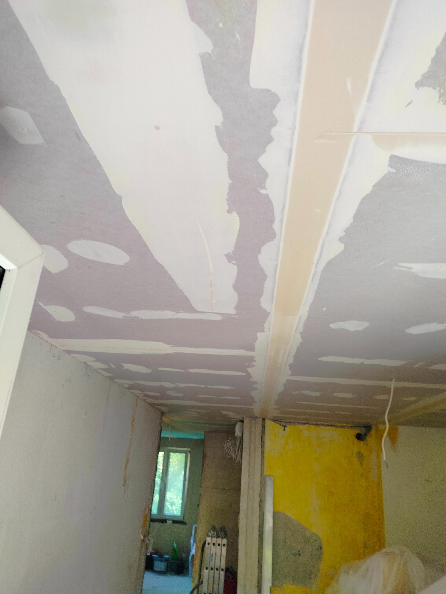 Niedokończony sufit z płyt gipsowo-kartonowych z widocznymi łączeniami i fragmentami gładzi, żółta ściana w tle, widok w głąb pomieszczenia z oknem.