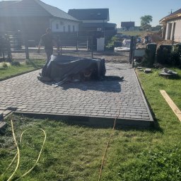 Serhii Molchanov Werolina - Ułożona szara kostka brukowa na podjeździe do domu, częściowo zakryta czarną folią, z widocznymi narzędziami i pracownikiem w tle, otoczona trawnikiem i ogrodzeniem.