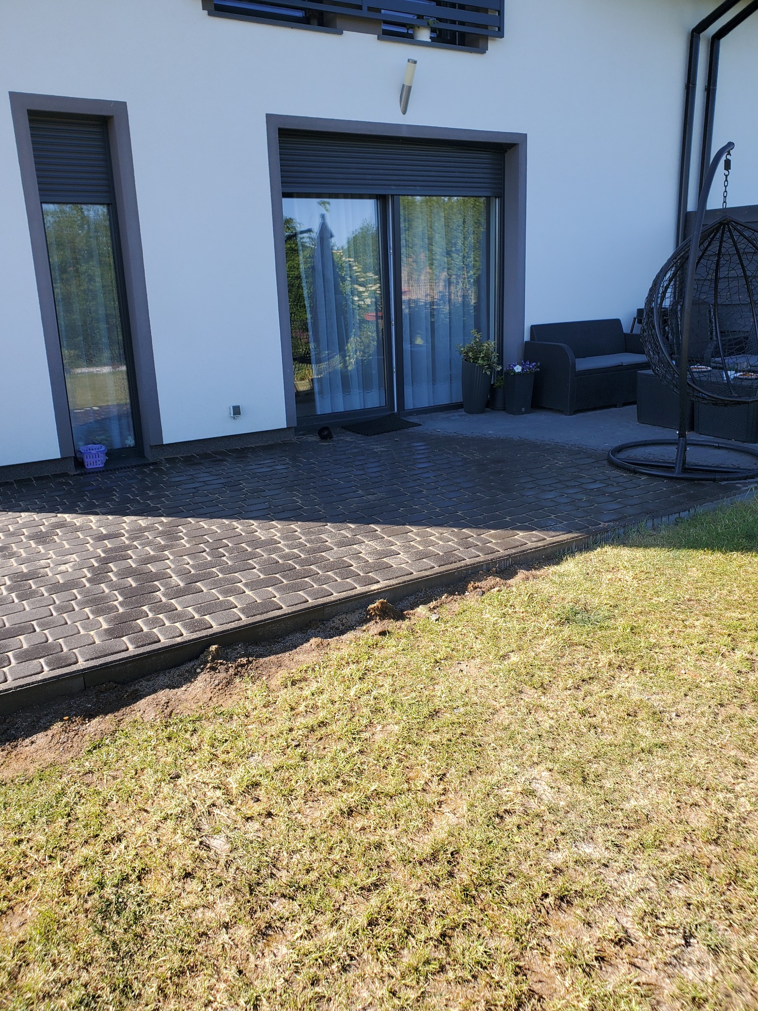 Ułożona, ciemnoszara kostka brukowa przed nowoczesnym domem z białymi ścianami i szarymi ramami okiennymi, widoczny fragment trawnika.