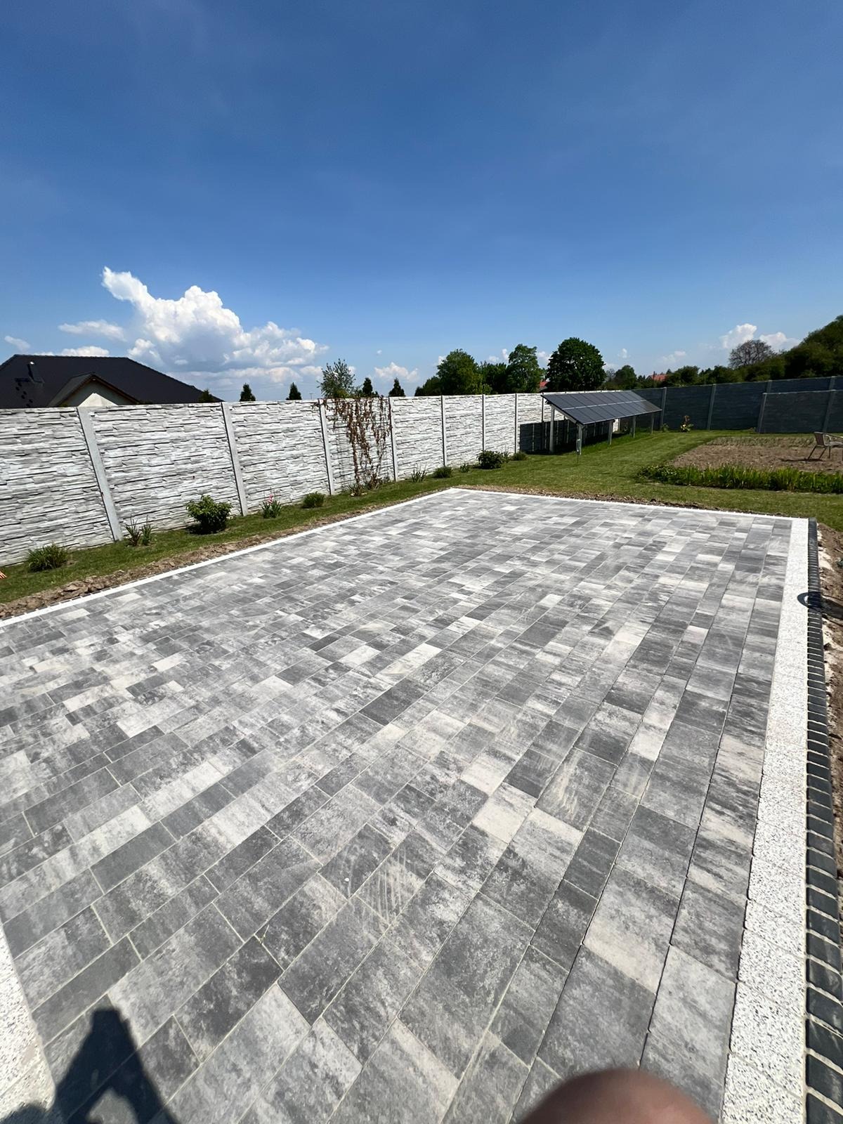 Ułożona kostka brukowa w odcieniach szarości na posesji z nowoczesnym ogrodzeniem z paneli imitujących kamień, panele solarne w tle.