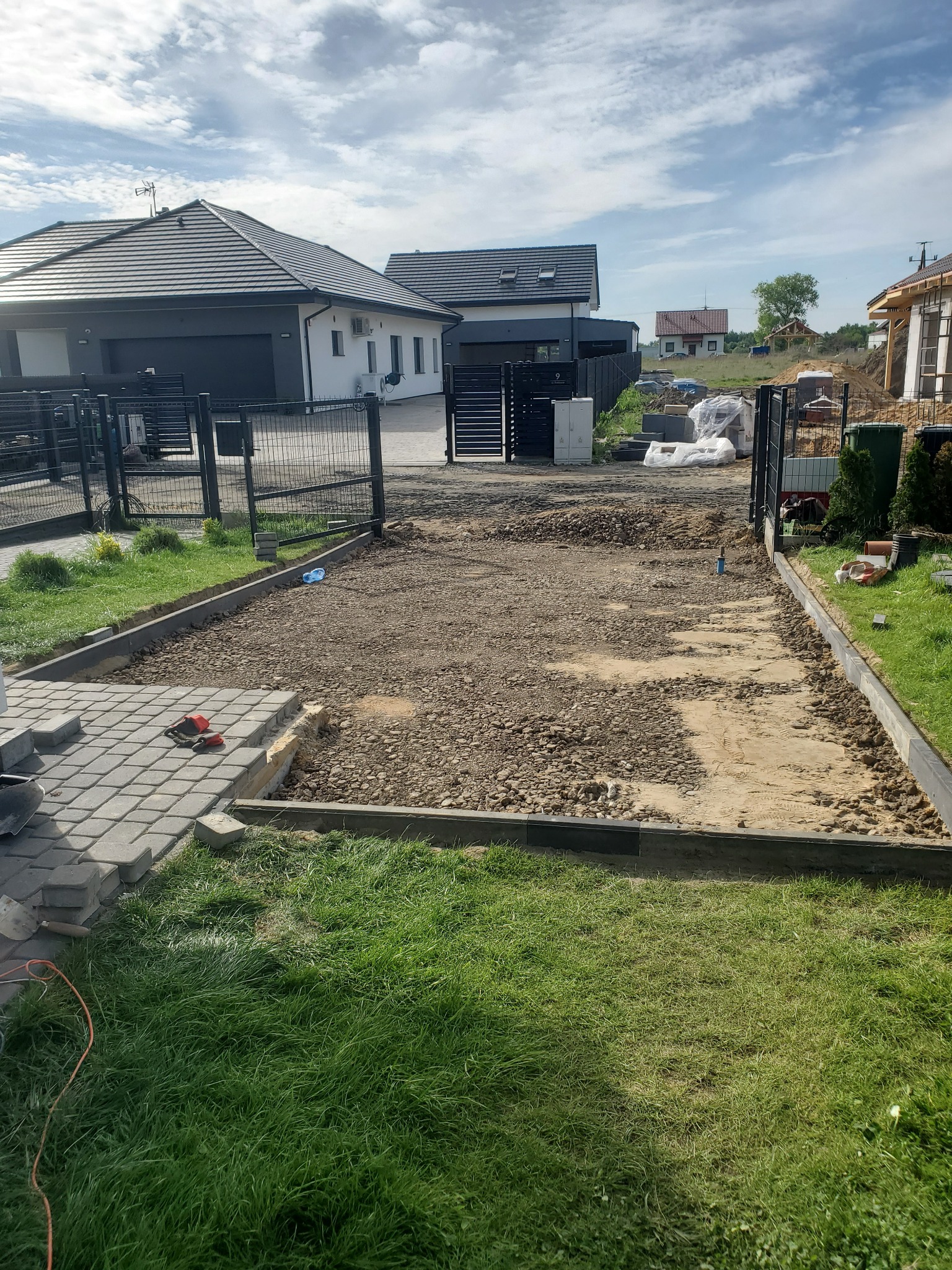 Teren przygotowany pod ułożenie kostki brukowej, widoczne obrzeża, podsypka, częściowo ułożona kostka, narzędzia i dom w tle.