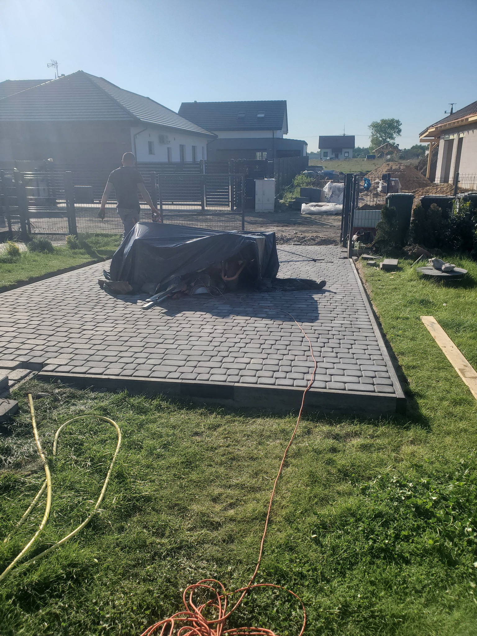 Ułożona szara kostka brukowa na podjeździe do domu, częściowo zakryta czarną folią, z widocznymi narzędziami i pracownikiem w tle, otoczona trawnikiem i ogrodzeniem.