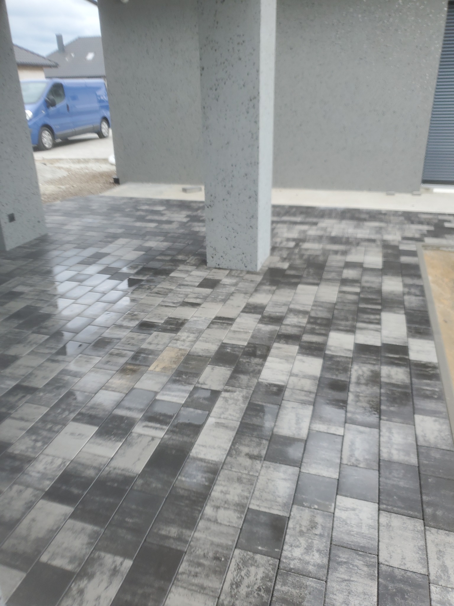 Świeżo ułożona, mokra kostka brukowa w odcieniach szarości na podjeździe, z widocznym fragmentem elewacji budynku i zaparkowanym niebieskim vanem w tle.