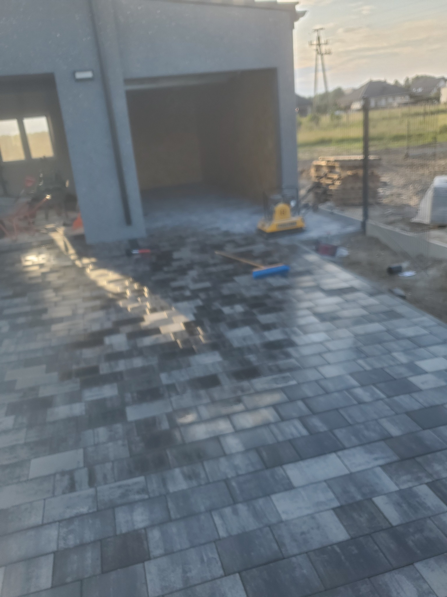 Świeżo ułożona, szara kostka brukowa przed garażem, z widoczną zagęszczarką i szczotką do zamiatania, w tle ogrodzenie z siatki i trawa.