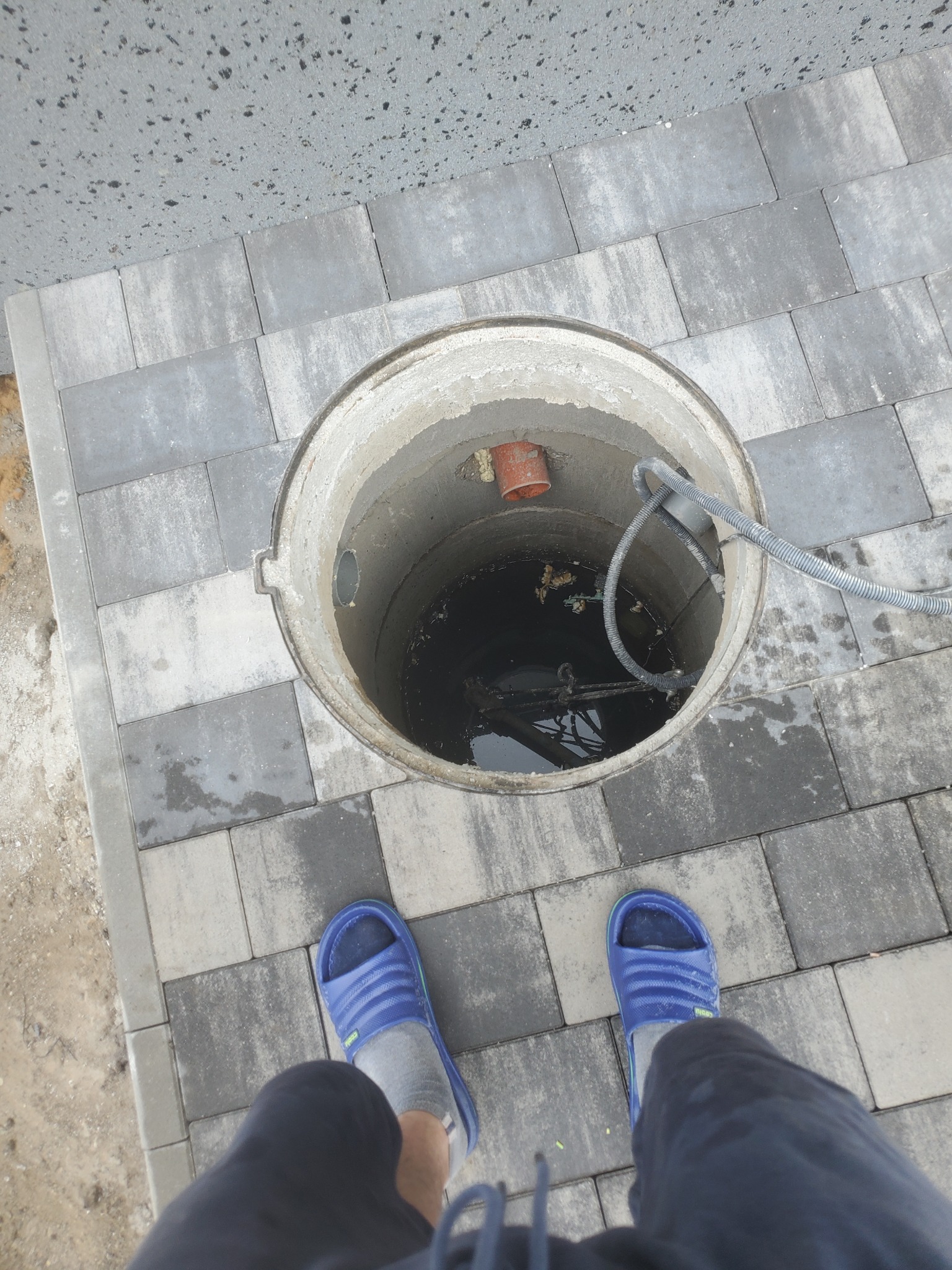 Studzienka kanalizacyjna z kablem i rurką wewnątrz, otoczona szarą kostką brukową, widok z góry z fragmentem osoby w niebieskich klapkach.