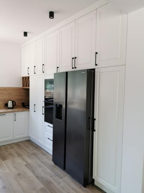 Biała kuchnia z zabudową od podłogi do sufitu, czarne, minimalistyczne uchwyty, duża, czarna lodówka side-by-side wkomponowana w zabudowę, drewniana podłoga i blat.