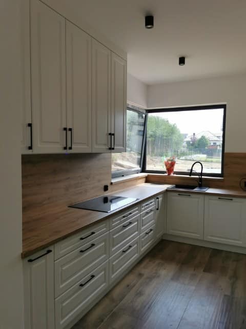 Jasna kuchnia z drewnianym blatem i czarnymi uchwytami, widok na okno z roślinami na parapecie.