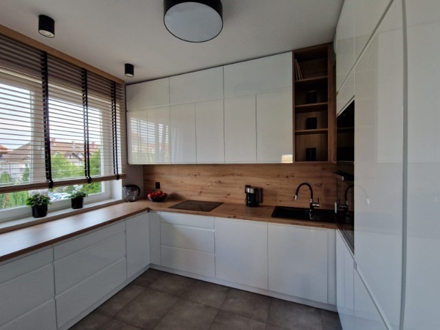 Biała kuchnia z frontami na wysoki połysk, drewnianym blatem i okładziną ścienną, czarną armaturą i żaluzjami drewnianymi w oknie.